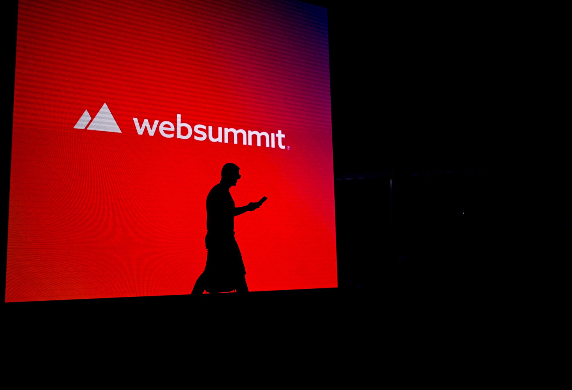 Silhueta de uma pessoa a caminhar e a mexer no telemóvel, à frente de um ecrã gigante com o logo da Web Summit.