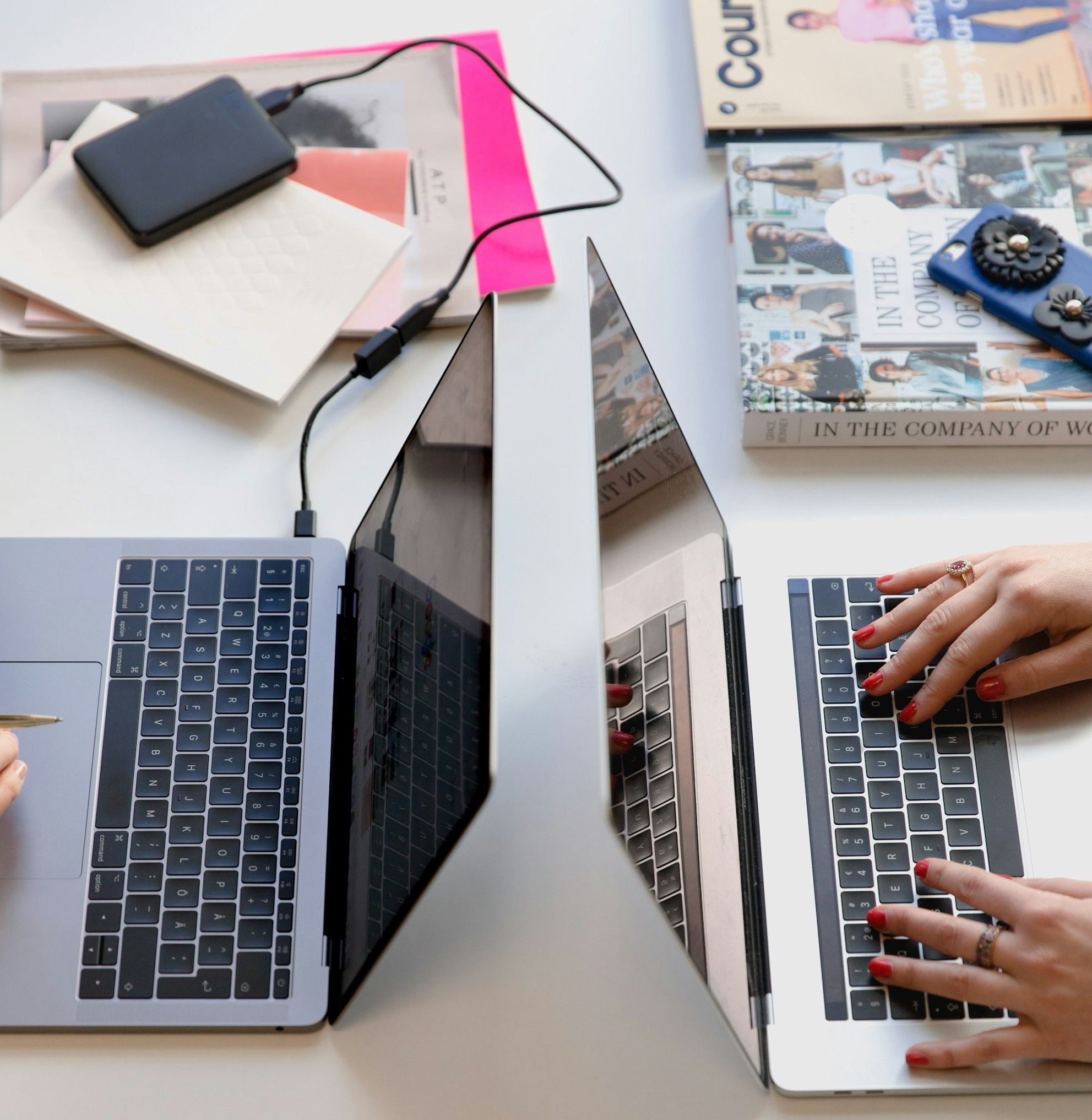 Dois MacBooks de costas um para o outro numa mesa com um disco externo, um iPhone e livros. Dos utilizadores, só as mãos estão visíveis.