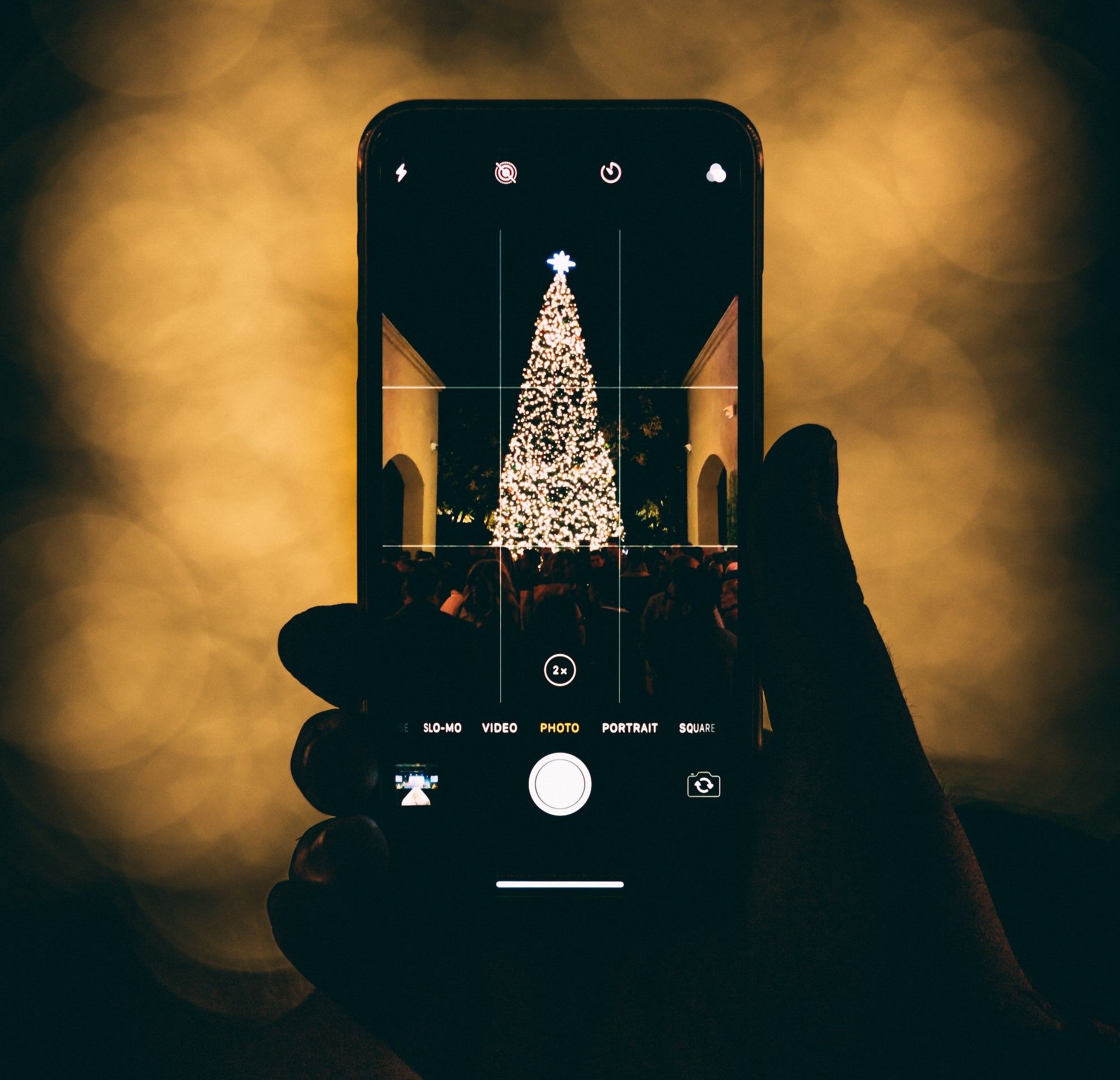 Mão direita a segurar um iPhone com a câmara aberta cujo ecrã mostra uma árvore de Natal bem iluminada entre dois arcos de pedra. À frente, um grande grupo de pessoas. 