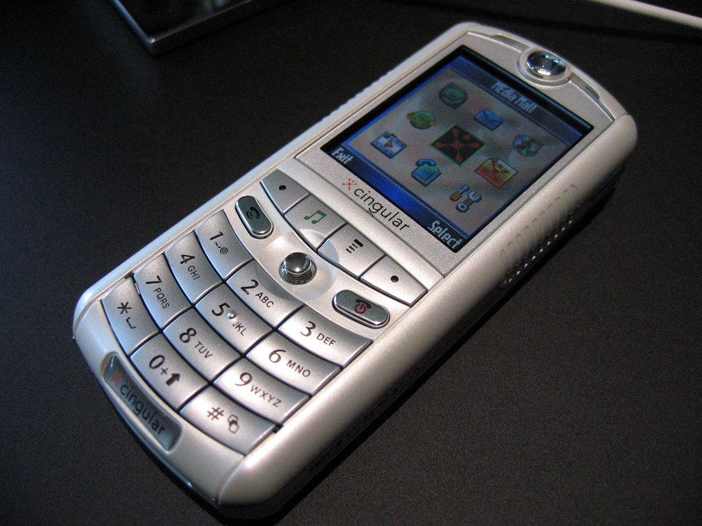 Motorola ROKR, na diagonal (com o teclado para a esquerda e para baixo) e com o ecrã ligado, pousado numa superfície.